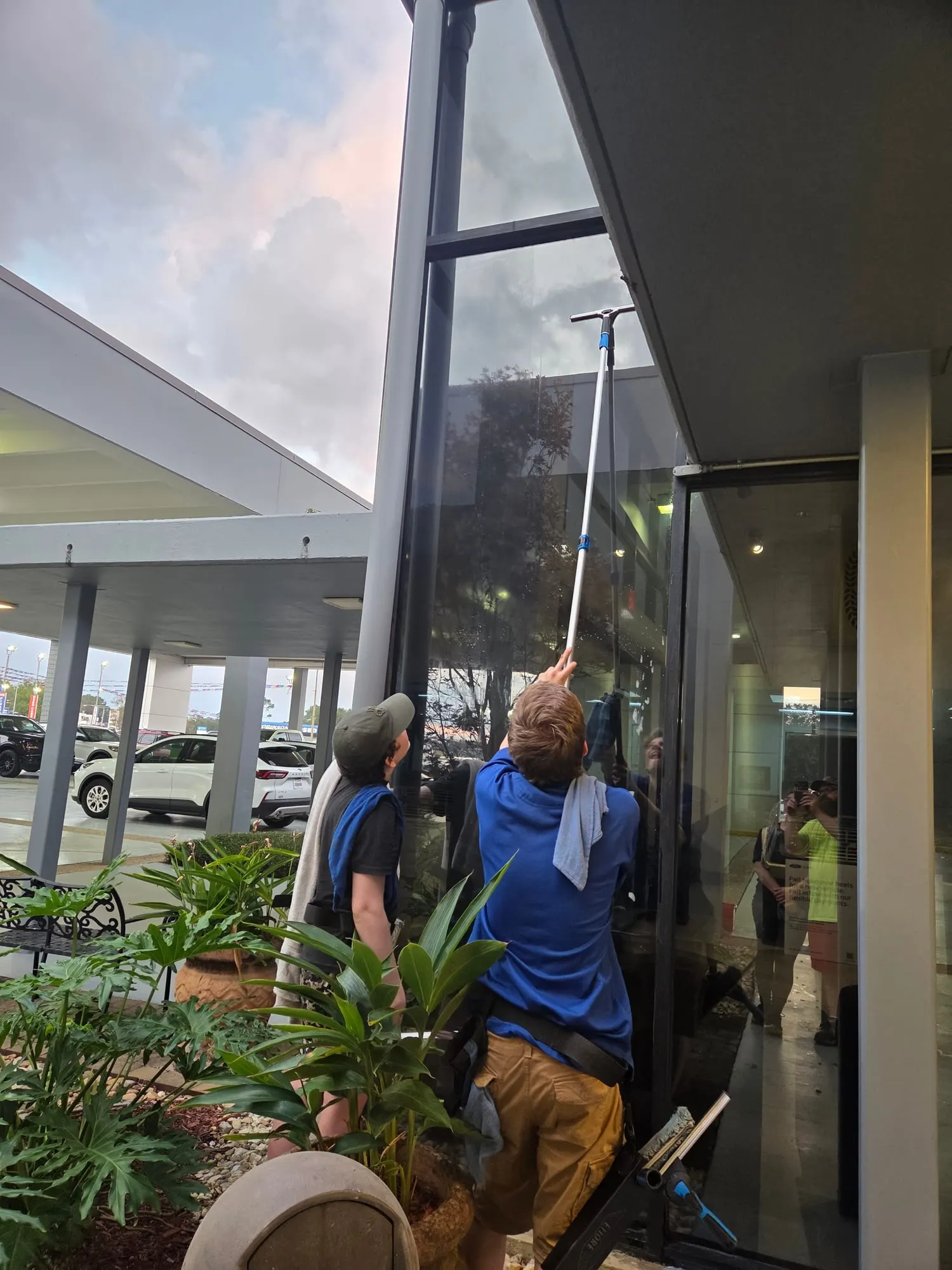 A two-person crew, seen from behind, cleaning tall commercial glass with a squeegee on an extension pole